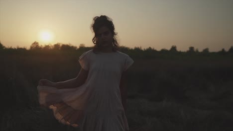 slow motion handheld shot of a young indian woman in a dress standing on a field during a beautiful sunset