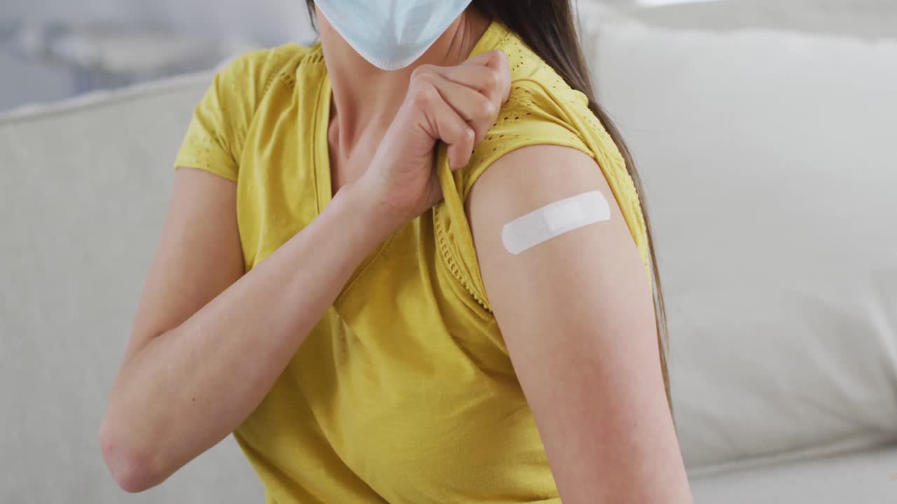 sección media de una mujer asiática con máscara facial sentada en el sofá mostrando el brazo con yeso después de la vacunación
