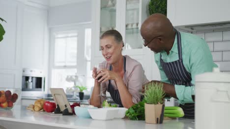 Sonriente-Pareja-Diversa-De-Ancianos-Usando-Delantales-Azules-Y-Bebiendo-Vino-En-La-Cocina