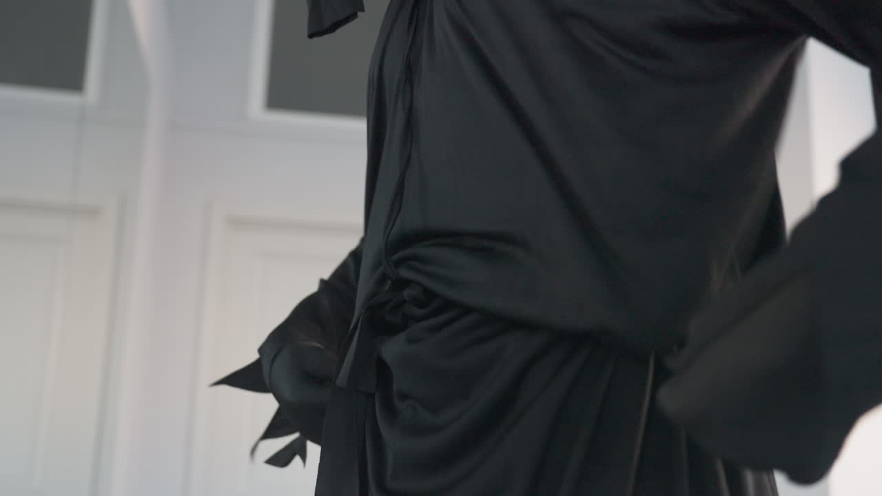 close up of person in black flowing outfit adjusting waist belt, detailed fabric folds visible, light gently reflecting off smooth dark costume, minimalist interior with soft background