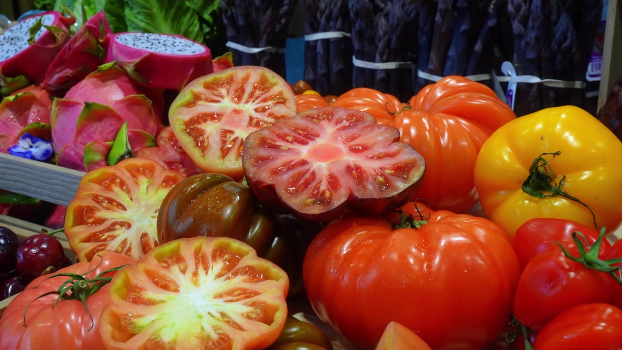 Heirloom-tomater hele og skåret i skiver på en udstilling af friske råvarer. Flerfarvede sorter med dragefrugt og asparges i baggrunden.