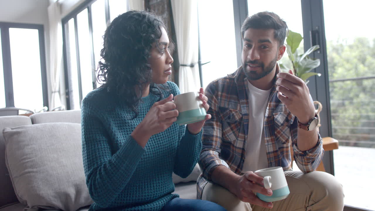 Happy biracial couple sitting on sofa, drinking coffee and talking at home, in slow motion