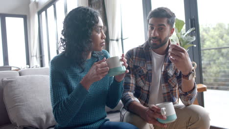 happy biracial couple sitting on sofa, drinking coffee and talking at home, in slow motion