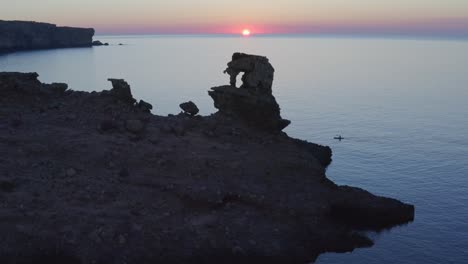 paddle boarder along the coast of menorca going past huge rocky cliff face