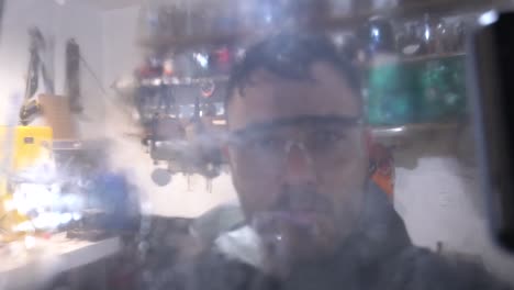 in a dimly-lit shed, a man sprays a tint onto a transparent glass, surrounded by a clutter of tools and equipment