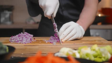 fresh onion being cut on a wooden board by young professional male chef in an elegant black shirt with tattoos and white gloves