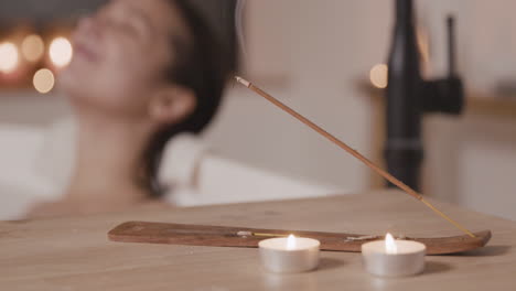 vue rapprochée d'une table avec des bougies et de l'encens, en arrière-plan une femme floue prenant un bain et buvant