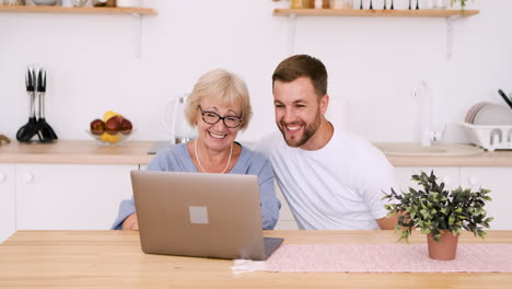 Glückliche-ältere-Mutter-Und-Erwachsener-Sohn-Sitzen-Am-Tisch-Im-Kitcken-Und-Sprechen-über-Videoanruf-Auf-Modernem-Laptop