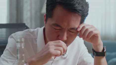 술에 취한 우울한 아시아 남자가 집에서 테이블을 부러뜨리는 주먹을 사용하기 전에 보드카를 마시고 있습니다.