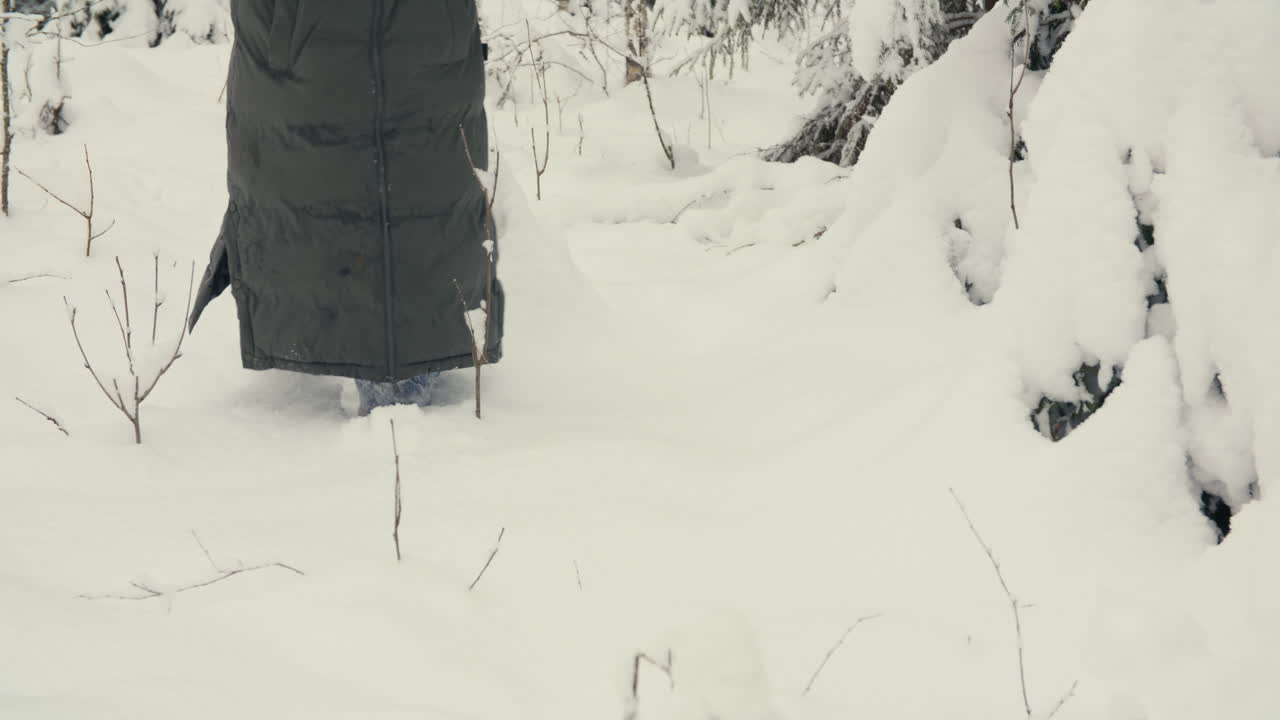 Primo piano posteriore di una persona che si muove nella neve profonda e intatta tra gli alberi, focalizzandosi sul movimento lento, sulla texture e sulle silenziose condizioni della foresta invernale.