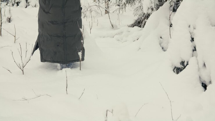 Primo piano posteriore di una persona che si muove nella neve profonda e intatta tra gli alberi, focalizzandosi sul movimento lento, sulla texture e sulle silenziose condizioni della foresta invernale.