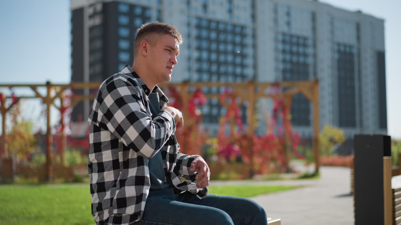 young student in checkered shirt sits on wooden bench reaching into pocket under bright sun with modern tall building and red blooming flowers in background