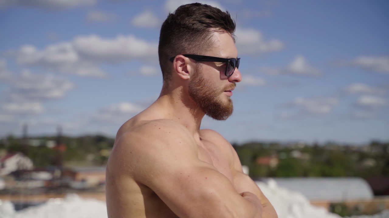 Muscular athlete on the top of the hill. Face of a bearded guy in black sunglasses outdoors on the blur city background. Close-up.