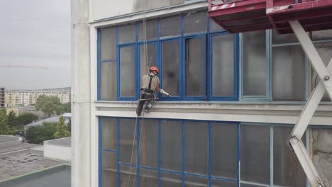 Toma-Aérea-Panorámica-A-Través-De-Un-Escalador-Industrial-Junto-A-Un-Andamio-Elevador-De-Tijera-Que-Trabaja-En-El-Sellado-De-Ventanas