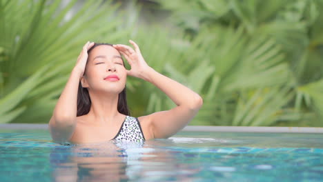 Porträt-Einer-Sexy-Exotischen-Frau,-Die-Ihr-Nasses-Haar-Im-Swimmingpool-Mit-Tropischer-Üppigkeit-Im-Hintergrund-Fixiert,-Vollbild-Zeitlupe