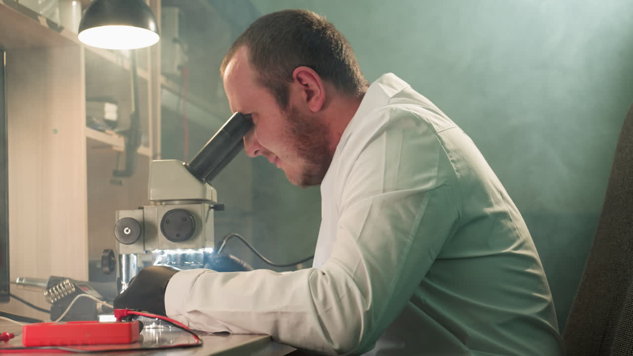una vista de cerca de un técnico enfocado en una bata de laboratorio blanca usando un microscopio para inspeccionar una placa de circuitos electrónicos, con una lámpara de escritorio encendida