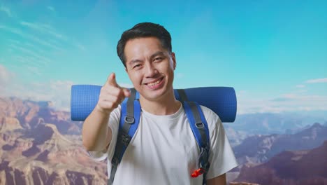 close up of asian male hiker with mountaineering backpack smiling, touching his chest, and pointing to camera while traveling at the top of mountain
