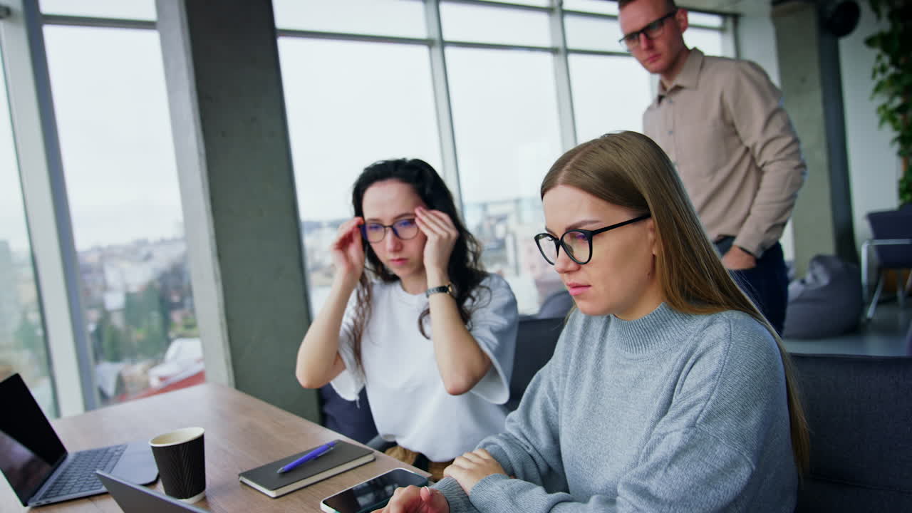 Female coworkers look at computer and seem to have some problem. Man walking behind them looks at screen and waves his head disapprovingly.