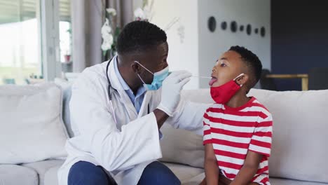 African-american-doctor-wearing-face-mask-taking-a-throat-swab-sample-of-boy-at-home