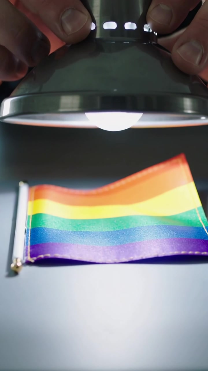Vertikal video: Mand griber metallampe, sænker lampen over et regnbueflag på et bord, oplyser farverne