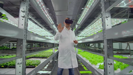 male farmer biologist in a white robe standing in the hallway of vertical farming with hydroponics with glasses virtualnoy reality around the green showcases with vegetables.