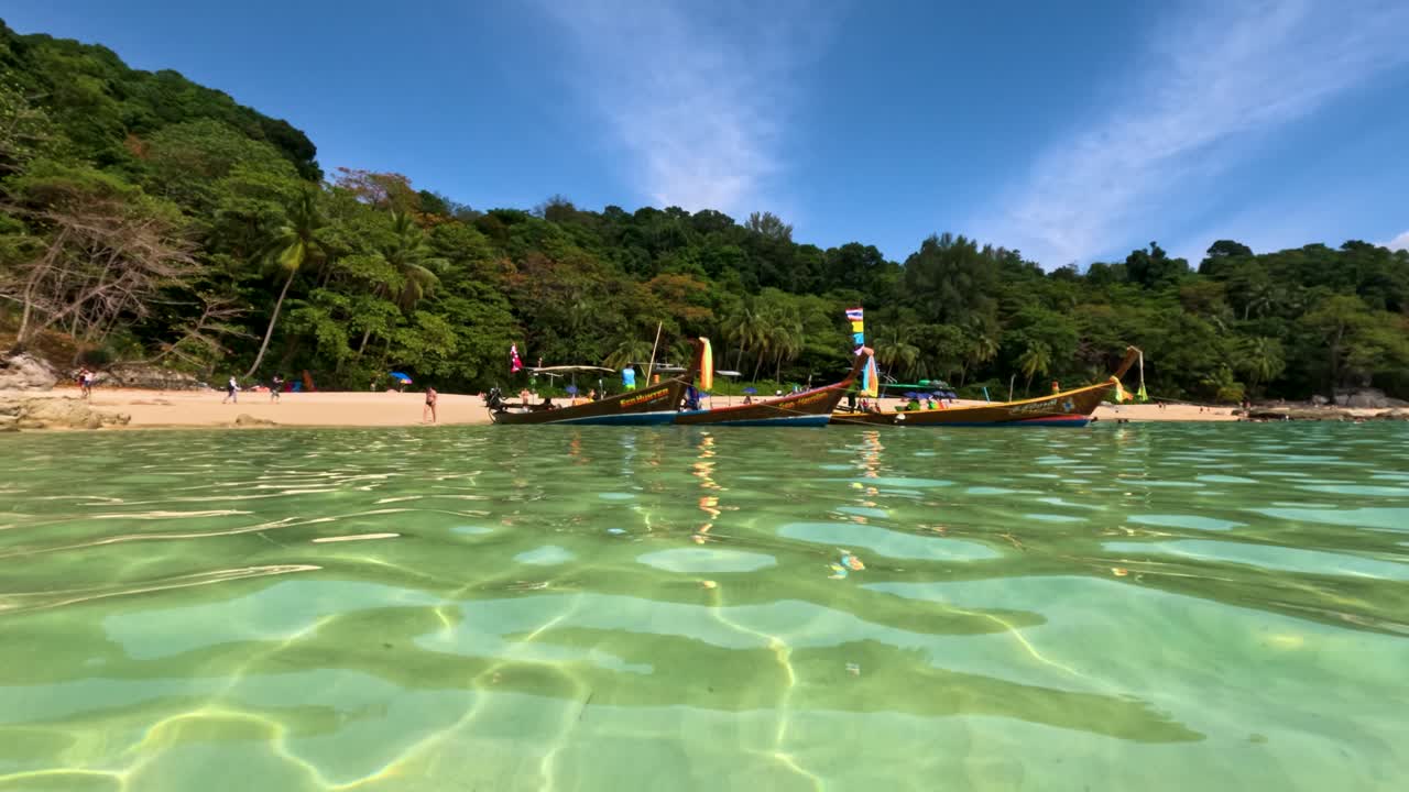 Barche longtail galleggiano su acque limpide e turchesi vicino a una spiaggia lussureggiante e tropicale a Phuket, Thailandia, in piena luce diurna.