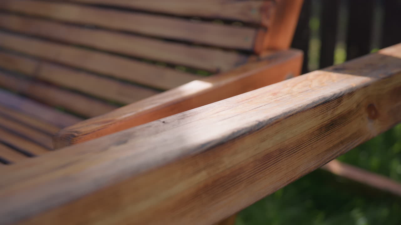 Close up of smooth wooden swing with metal chain gently swaying in quiet backyard, sun illuminating warm wood tones and blurred fence in background, evoking calm, relaxation, and rustic charm
