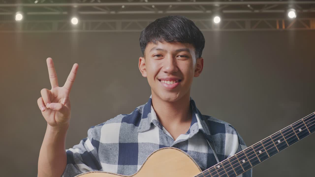 기타를 들고 있는 아시아 십대가 무대에서 평화와 미소를 짓는 몸짓을 보여준다.