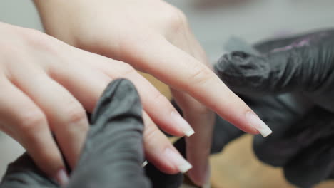 close up view of nail technician wearing black gloves inspecting fingernail length and shape of customer s hand using metal file during detailed manicure session in professional salon setting
