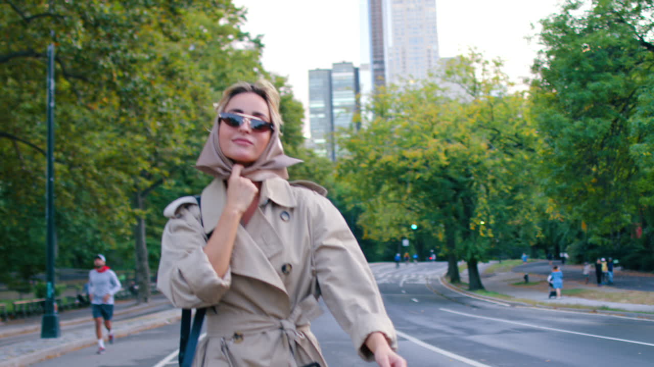 New York, USA, 2 aprile 2026: Donna con occhiali da sole e foulard che cammina su una strada cittadina. Mezza figura di una donna in trench e foulard che cammina verso la telecamera in un parco urbano di New York