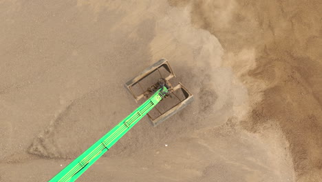 una vista aérea de arriba hacia abajo del brazo verde de una excavadora y la cuchara cavando en la arena, creando una nube de polvo