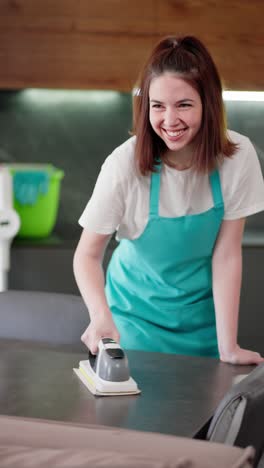 vertical video of a confident brunette cleaning lady in a blue apron polishing a glossy table in a modern apartment. portrait of a confident brunette girl a professional cleaner who does cleaning on call