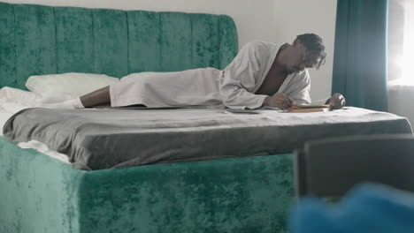 Man in white bathrobe laying on bed, focused on reading book, flipping through pages with one hand, green velvet headboard in background, soft light coming from window, peaceful indoor atmosphere
