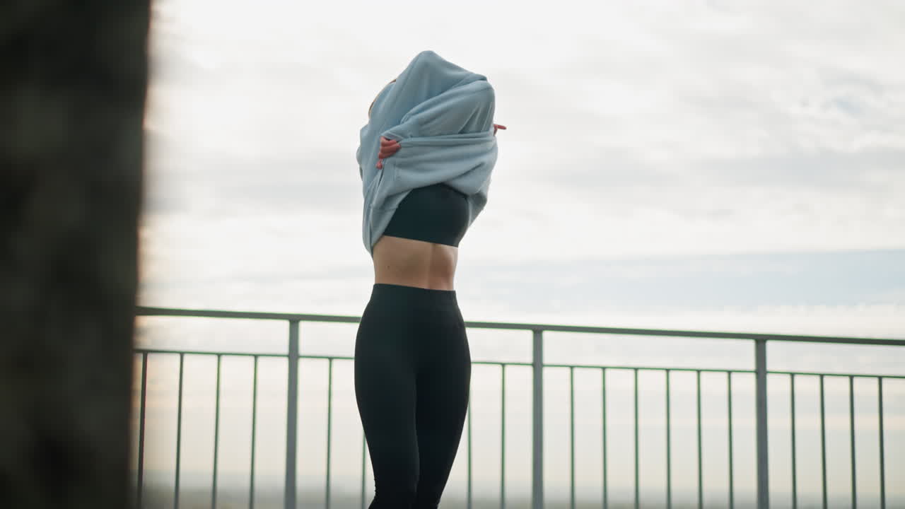 Girl removing hoodie, leaving just her singlet and leggings, standing in front of an iron fence, background features a calm outdoor setting, perfect for fitness, exercise, and casual lifestyle