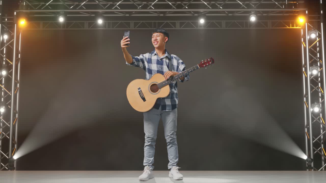 cuerpo lleno de adolescente asiático con la mano agitando la guitarra y teniendo una llamada de video en el teléfono inteligente mientras está de pie en el escenario