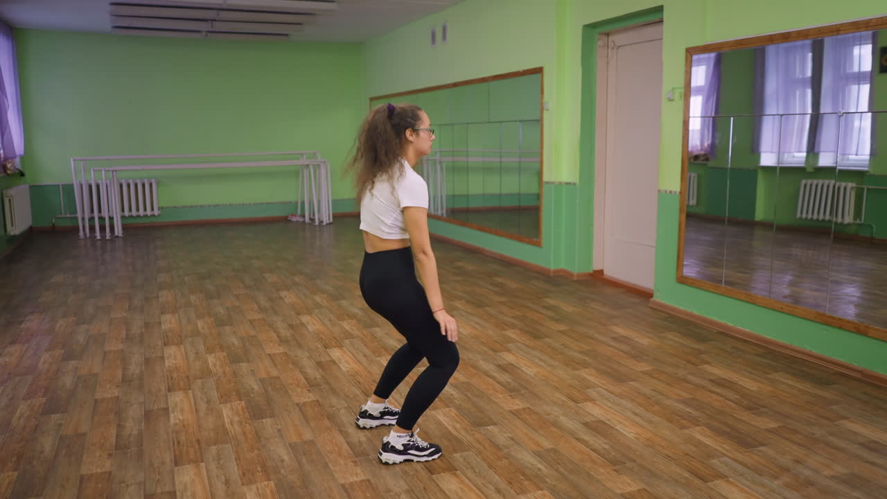 Beyaz üst ve siyah tayt giyen güzel bir kadın, parlak yeşil bir stüdyoda aynanın önünde saçlarını savurarak dans ediyor, ahşap zeminde spor ayakkabıları, enerjik hareket, eğlenceli ritim, kendinden emin poz