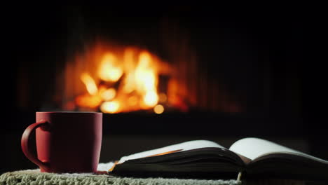 an open book and a cup of hot tea on the background of the fireplace where the fire is burning