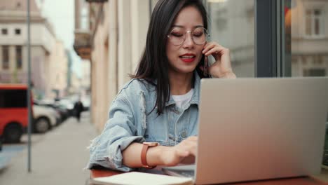 Junges-Schönes-Junges-Mädchen-Mit-Brille,-Das-Am-Laptop-Auf-Der-Caféterrasse-In-Der-Stadt-Arbeitet-Und-Mit-Dem-Handy-Spricht