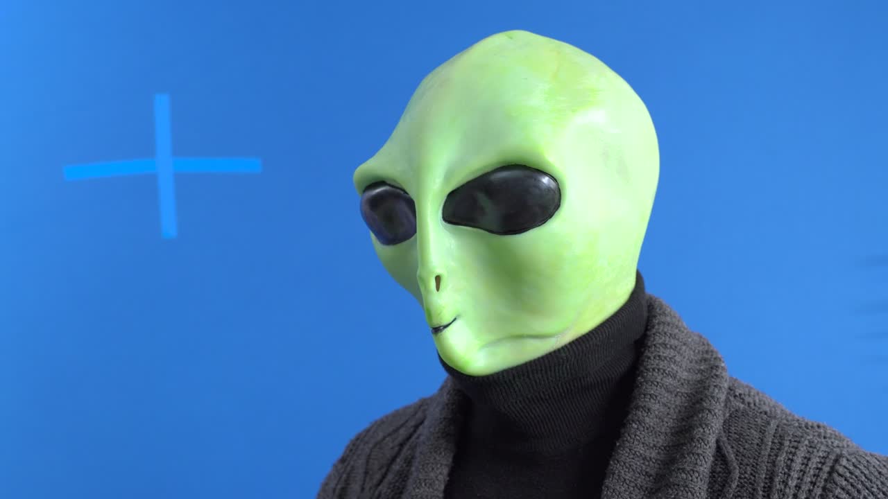 hombre en traje alienígena y chaqueta de punto cálida bebiendo amablemente bebida caliente de una taza temática con su dedo meñique sobresaliendo contra el fondo azul chromakey