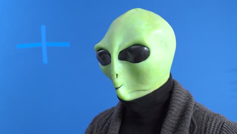 man in alien costume and warm knitted jacket mannerly drinks hot drink from themed mug with his little finger sticking out against blue chromakey background