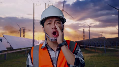 close up of asian male engineer with safety helmet yelling with hand over mouth while standing with solar panel and wind turbines