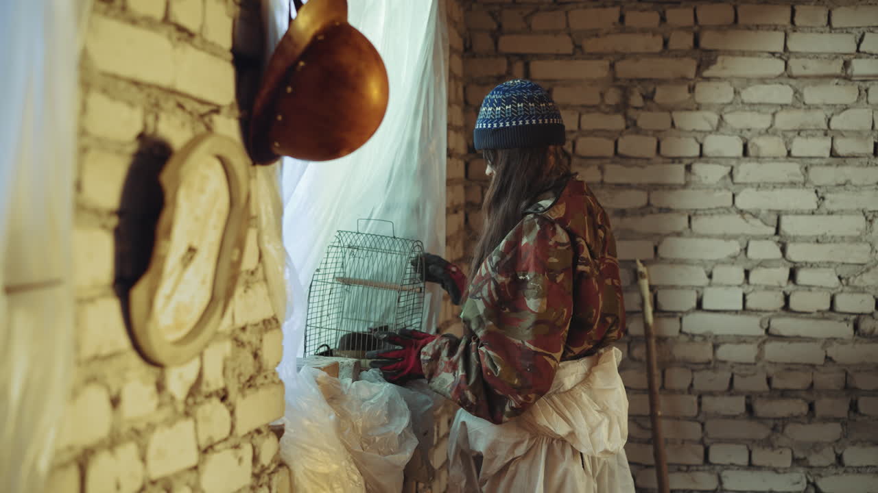 Homeless kid in military clothes carefully placing rat inside old metal cage by window with soft daylight through curtain, symbolizing survival and tense atmosphere in abandoned shelter environment