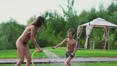 mom and son playing on the lawn pouring water laughing and having fun on the playground with a lawn on the background of his house near the lake