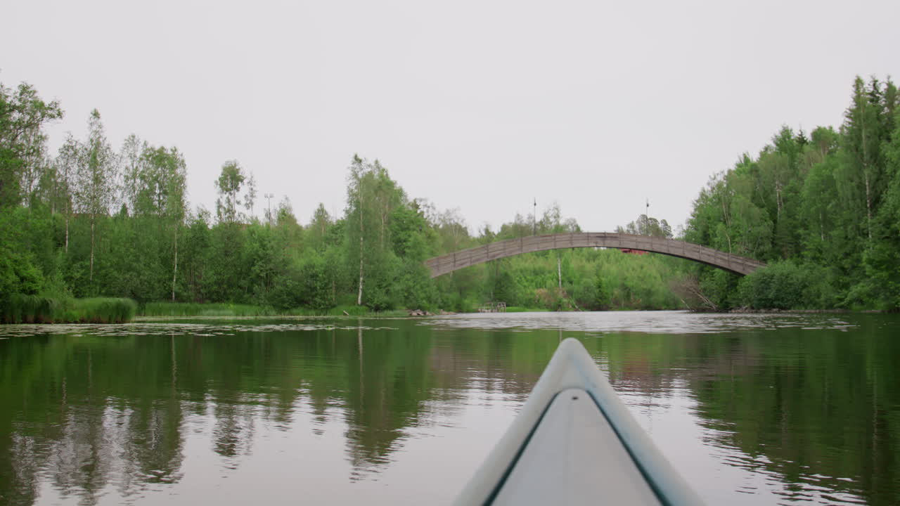 Point of view from canoe gliding on calm river towards rustic wooden footbridge, surrounded by dense green trees, tranquil water reflections and overcast daylight in eastern Norway