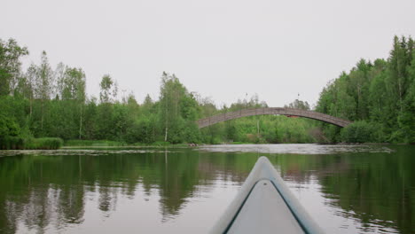 point of view from canoe gliding on calm river towards rustic wooden footbridge, surrounded by dense green trees, tranquil water reflections and overcast daylight in eastern norway