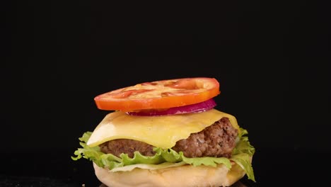 a sesame seed bun top is dropped onto a stacked beef burger with lettuce, cheese, tomato, and onion against a black background, using bright studio lighting and slow-motion capture