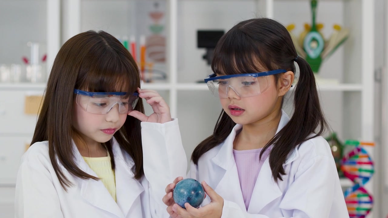 Siswi-siswi Asia mengenakan kacamata pengaman mengamati model planet di laboratorium sains yang terang.