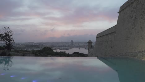 Vista-Nublada-Y-Colorida-De-La-Puesta-De-Sol-Sobre-La-Vista-Del-Gran-Puerto-De-Valletta-Desde-La-Piscina-Infinita-De-Lujo---Toma-De-Cámara-Lenta-Deslizante