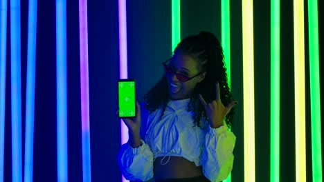 portrait of a young lovely female african american in stylish sunglasses points a finger at a smartphone with a green screen chroma key and making a rock gesture. slow motion