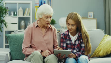 Pretty-Teenager-Girl-Sitting-On-The-Couch-In-The-Nice-Room,-Using-Tablet-Computer-And-Talking-With-Her-Grandmother-Who-Sitting-Next-To-Her